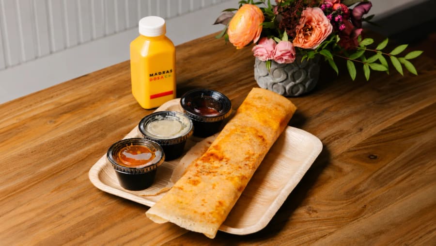 A long golden dosa on a tray with three dipping sauces and a Madras Dosa Co bottled drink