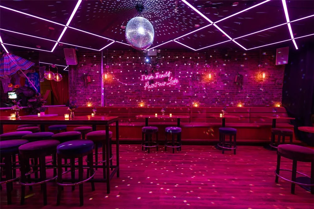 Pink-lit event room at The Factory 380 with a disco ball, neon sign, banquette seating and high-top tables